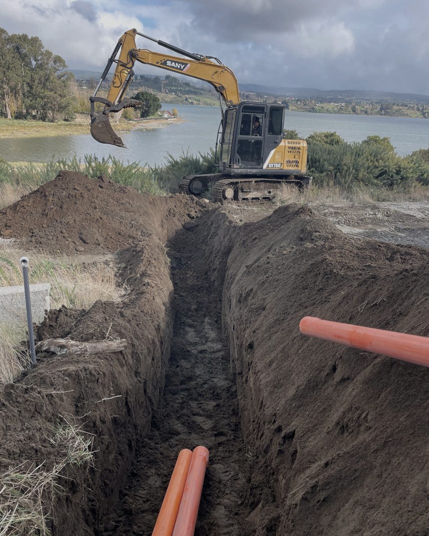 Zanja de canalización subterránea con ductos y retroexcavadora junto a un cuerpo de agua en Chiloé
